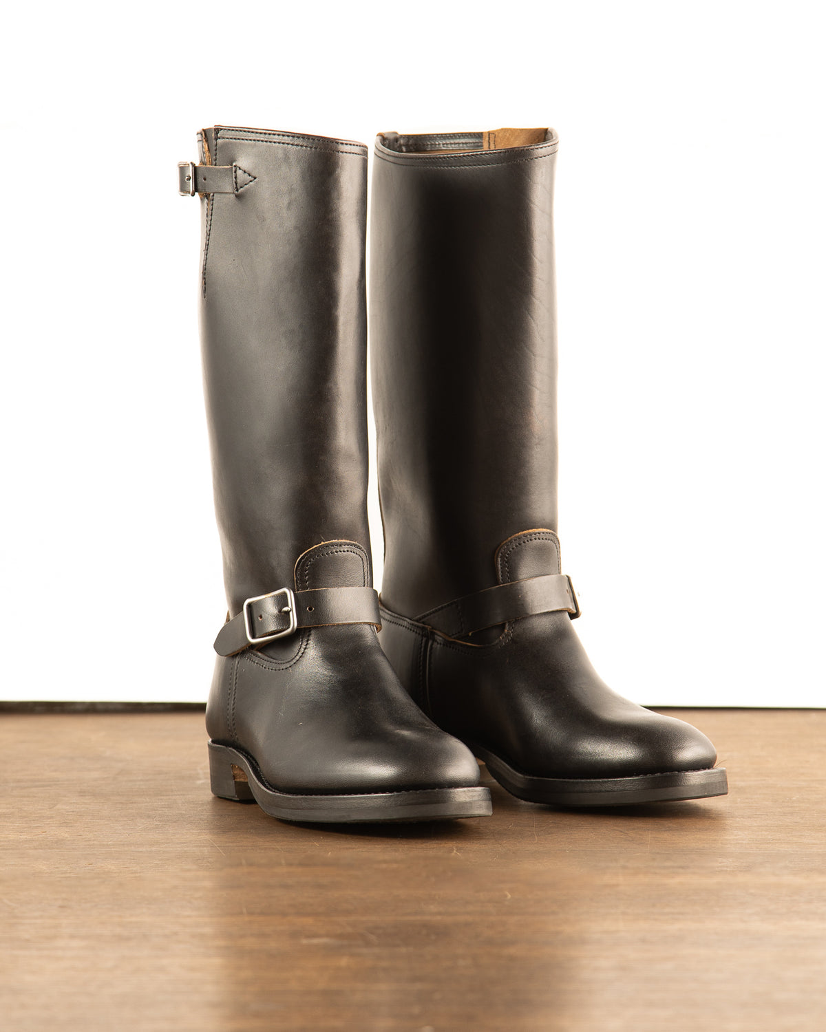 Black full-grain tall shaft aviator engineer boots with single ankle buckle strap and gunmetal silver rectangular hardware on wood floor