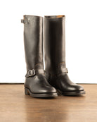 Black full-grain tall shaft aviator engineer boots with single ankle buckle strap and gunmetal silver rectangular hardware on wood floor