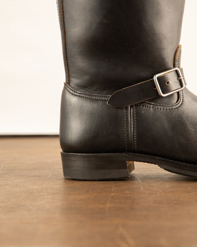 Close-up of a black leather boot with a buckle on a wooden floor.